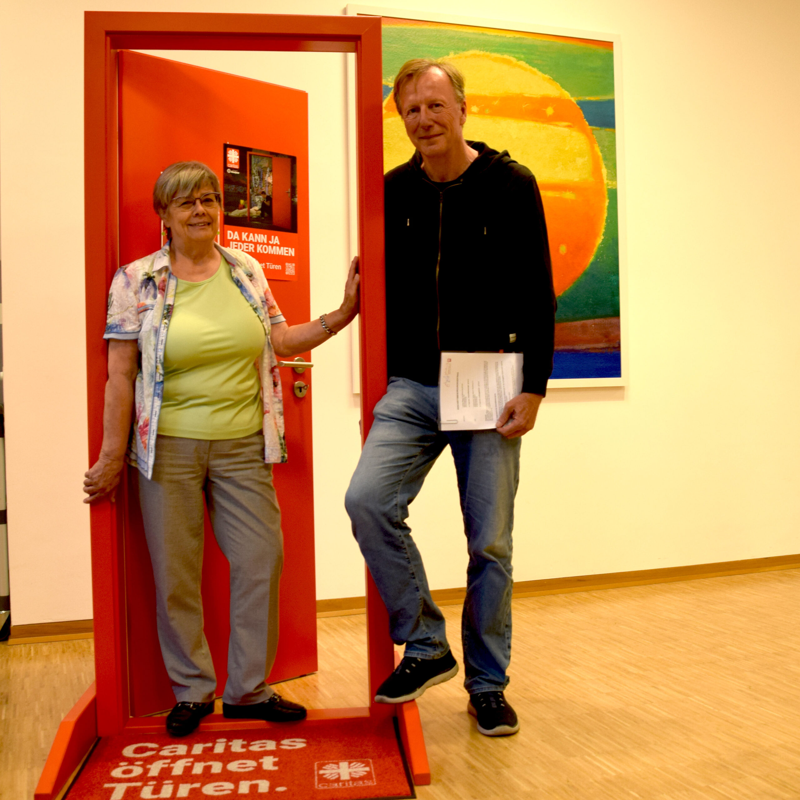 Rosemarie Horlemann und Stefan Weber stehen mit dem unterschriebenen Geschäftsbesorgungsvertrag in der roten Caritastüre.